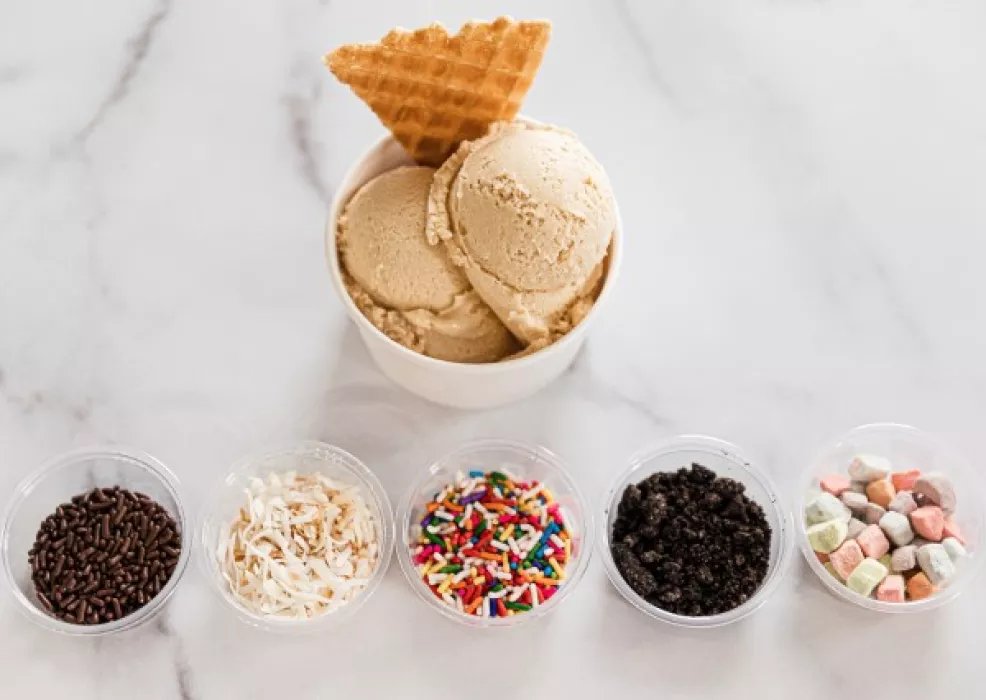 gelato scoops in a cup with a waffle wedge and 5 containers of toppings lined up below it